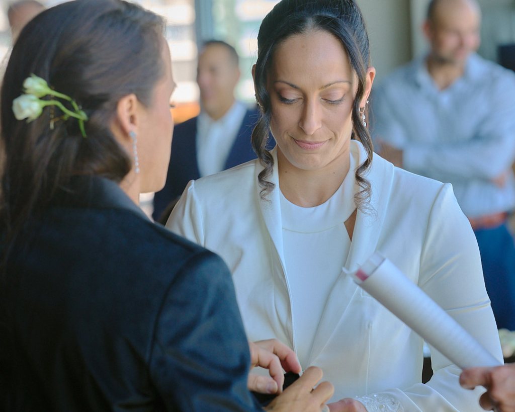 A close-up of a woman in a white outfit smiling at another woman with long hair and a flower, both engaged in a heartfelt conversation.